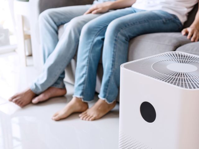 Close up of white air purifier on the floor in living room the background of young Asian couple relaxes on sofa. Concept of caring for fresh air at home. Caring for well-being and health.